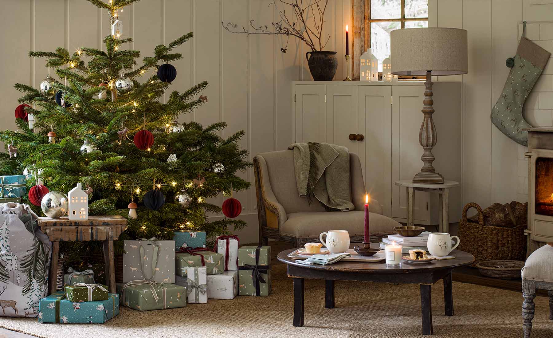 A Christmas tree with beautiful presents sat underneath, an armchair sits in the corner with a coffee table place in front with Christmas tableware by Sophie Allport,
