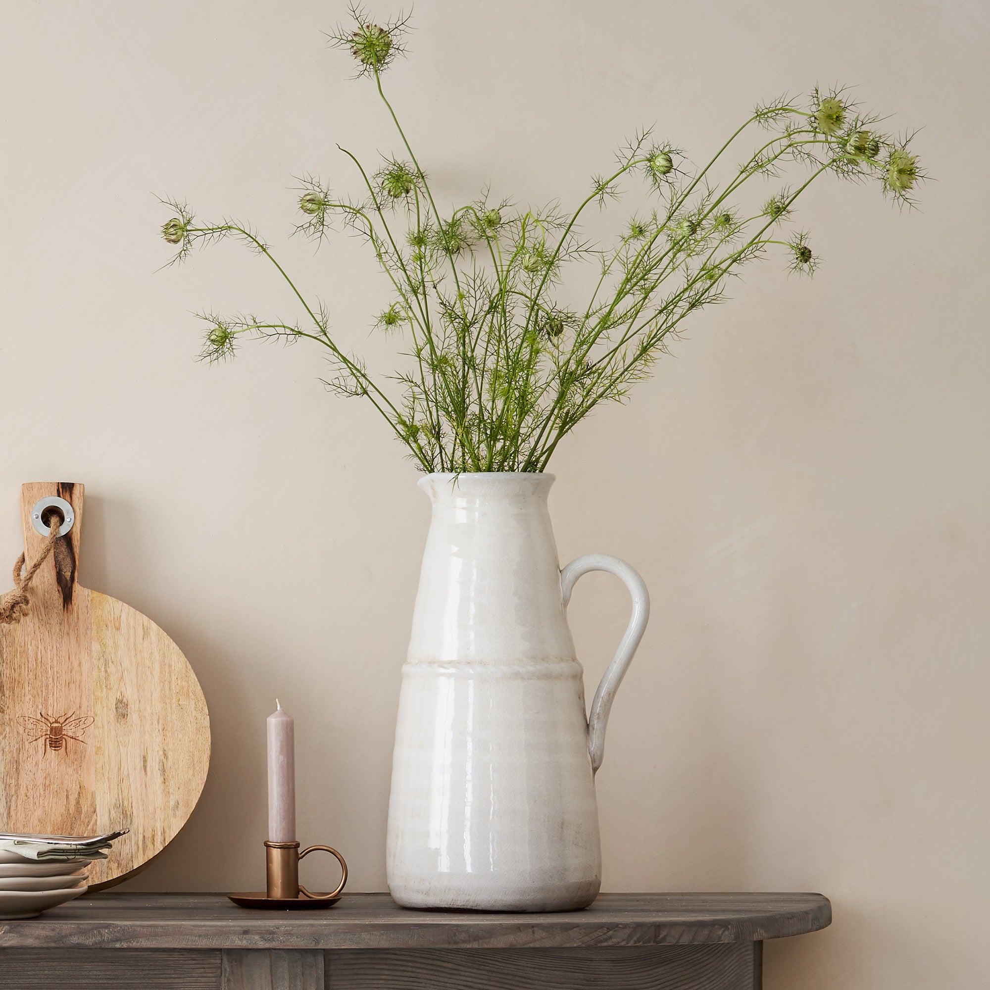 White ceramic pitcher with greenery on a wooden surface against a beige wall